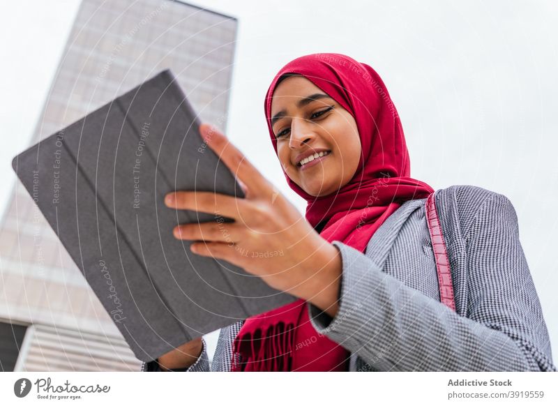 Positive ethnic woman in headscarf using tablet on street browsing hijab muslim city smile surfing female arab young red color device gadget connection rest