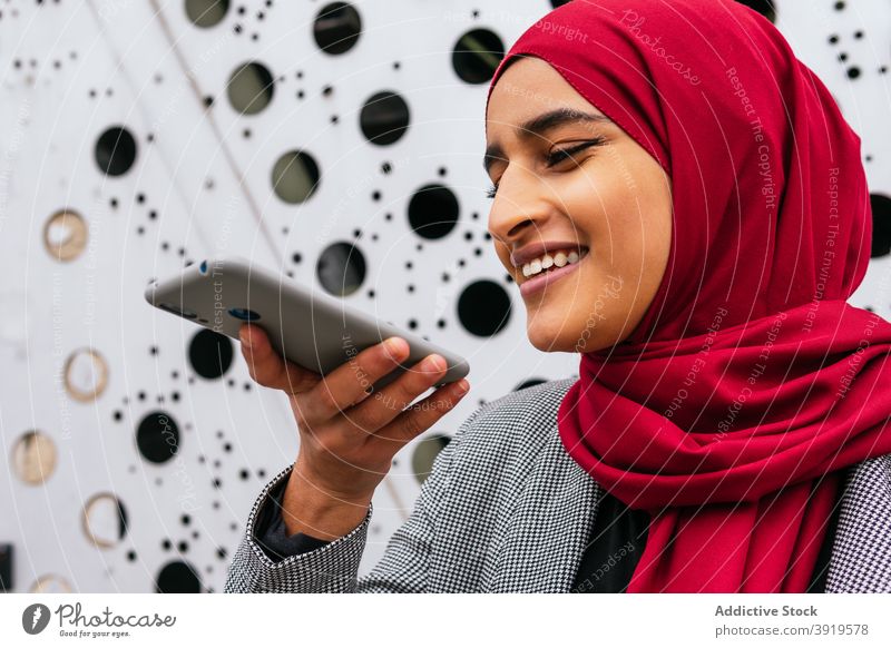 Delighted ethnic woman recording audio message on smartphone voice communicate social media hijab headscarf female arab street smile optimist internet