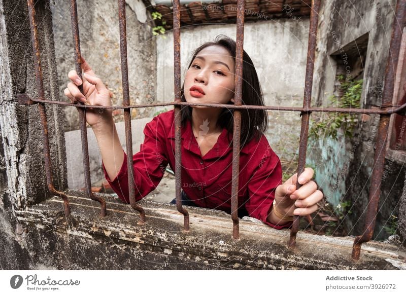 Ethnic woman looking through window with metal grid in old house abandoned weathered concept restriction shabby grunge dramatic confinement young asian ethnic