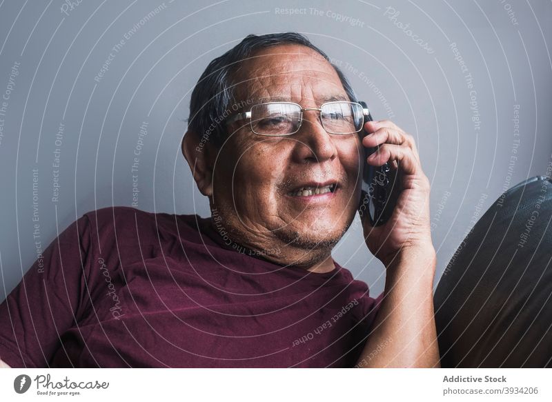 Ethnic senior man talking on smartphone at home speak elderly mobile sofa male ethnic call couch pensioner retire connection device gadget appearance wrinkle