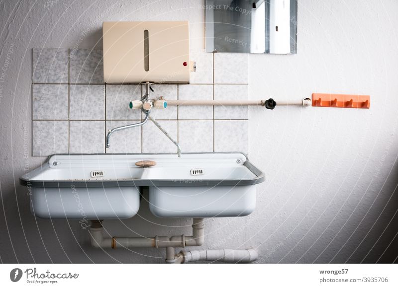Poor furnishings | one double sink, one bar of soap, one hot water heater on a white wall with a small tile backsplash, one rectangular mirror and one orange hook rail