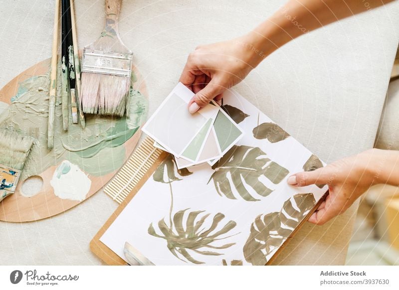 Crop artist with paper color palette and painting in workshop pastel tone monstera sample craftswoman female artisan green table studio creative hobby handmade