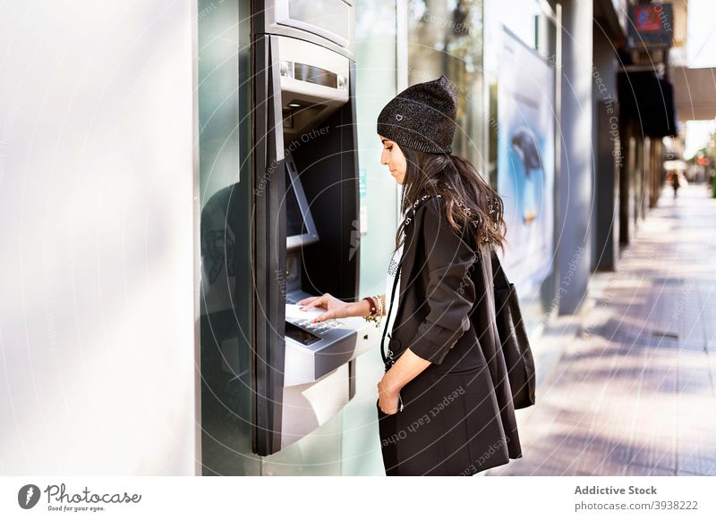 Concentrated young ethnic woman withdrawing money from ATM machine using atm cash bank pin city street style female trendy casual hat credit debit automatic