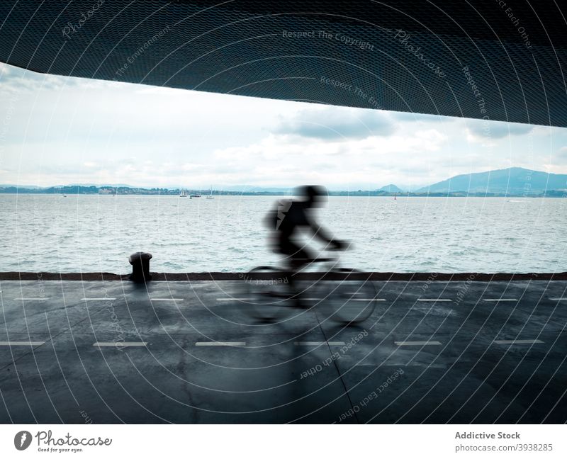 Unrecognizable man riding bike along embankment at seafront on cloudy day ride bicycle road nature activity speed motion male helmet vehicle transport coast