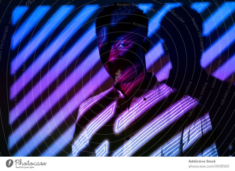 Black man in neon lights in studio illuminate stripe shape color trendy vogue male black ethnic african american dark style fashion individuality vivid serious