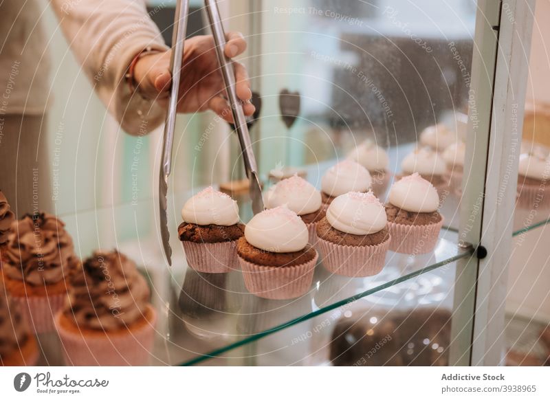 Seller taking cupcake from display in confectionery shop sweet seller take candy dessert cream tong female retail choice store select pick food bakery pastry