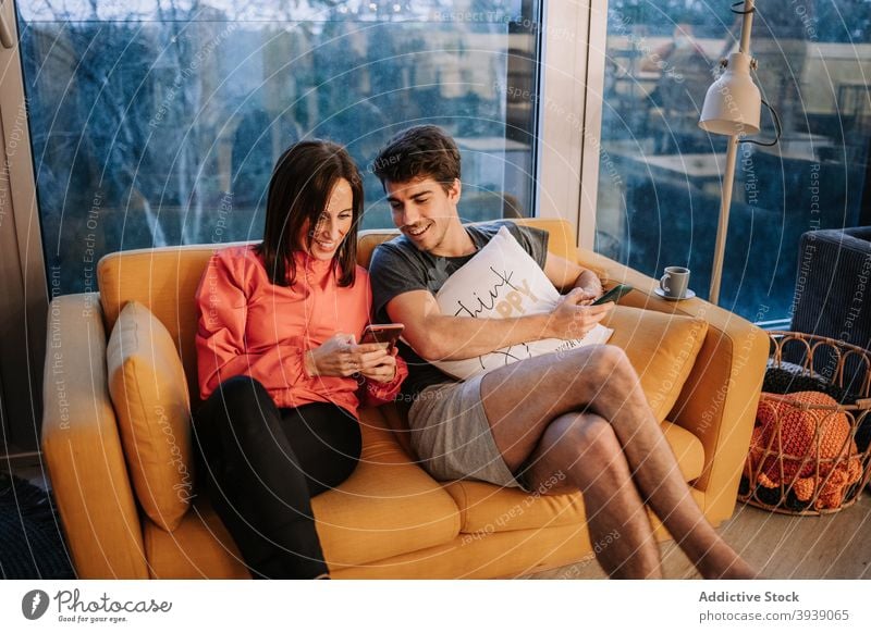 Smiling friends sitting on sofa and using smartphone together relax home weekend watch video mobile online internet connection gadget device enjoy communicate