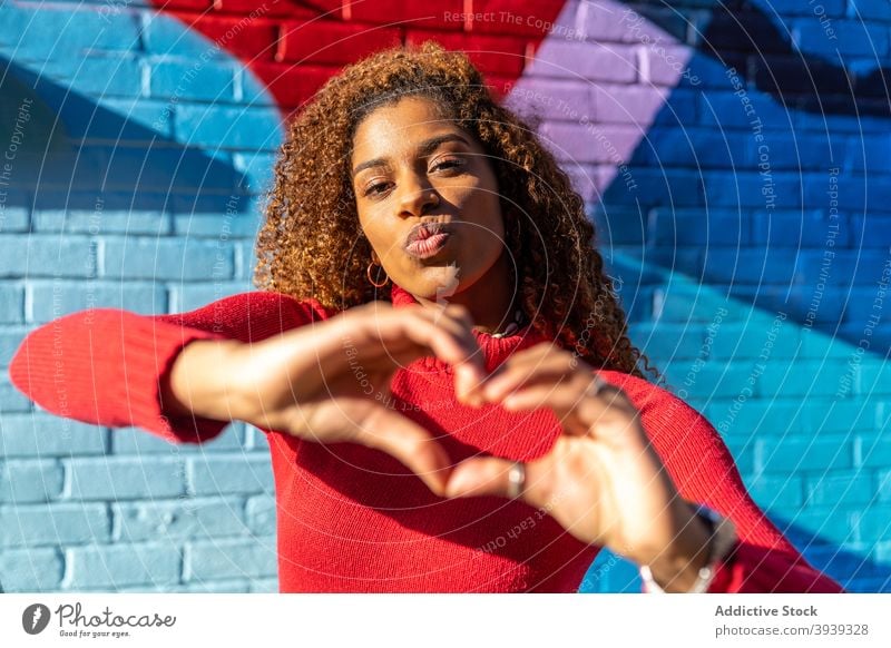 Content young black woman pouting lips and gesturing heart near graffiti wall gesture style confident street street art cool appearance happy female ethnic