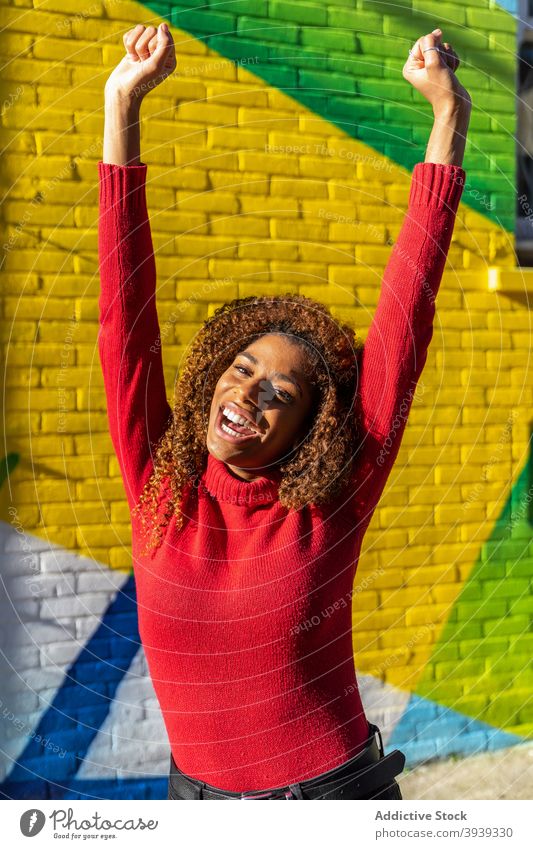 Satisfied young black woman clenching fists with raised arms against graffiti wall excited win satisfied arms raised fist up happy mouth opened clench fist