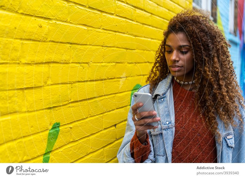 Confident young black woman using mobile phone and walking along street near multicolored building smartphone graffiti colorful pedestrian communicate content