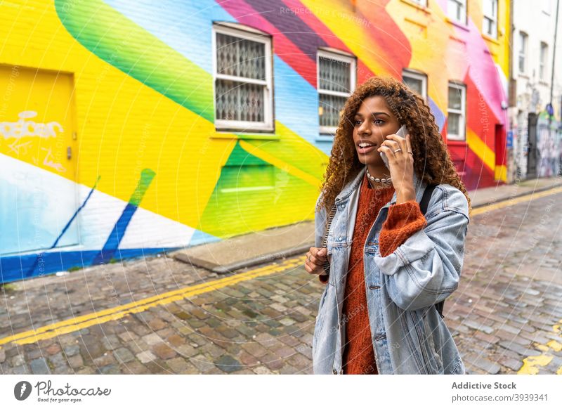 Confident young black woman talking on smartphone and walking along street near multicolored building graffiti colorful phone call pedestrian conversation
