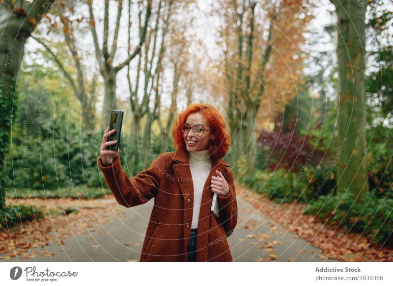 Positive woman using mobile phone in park speak smartphone redhead red hair cheerful enjoy conversation discuss female alley coat autumn gadget happy device