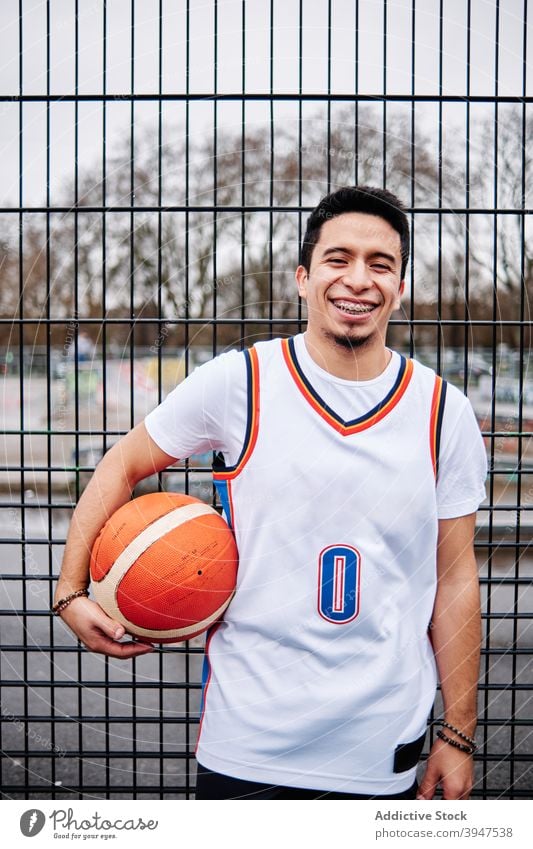 Latino guy posing with a basketball by the fence of a court outdoors. Sport concept. laughing friendly player individuality laughter motivation proud