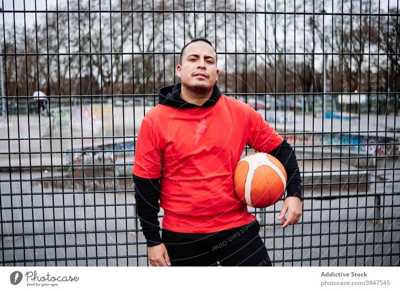 Portrait of latino guy posing with a basketball, looking at camera by the fence of a court. player horizontal attitude individuality pride vitality aspirations