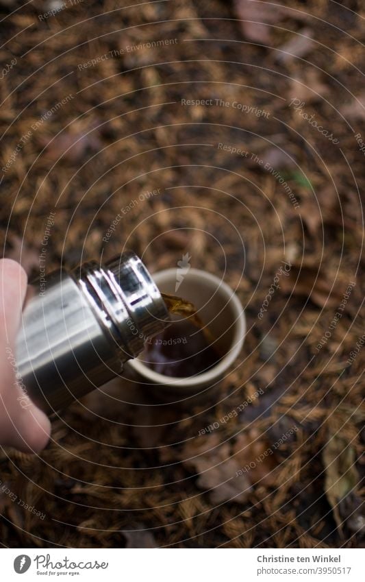 Break in the forest. A cup stands on the forest floor, this is covered with brown autumn leaves. Tea / coffee is poured from a silvery thermos bottle. tea break