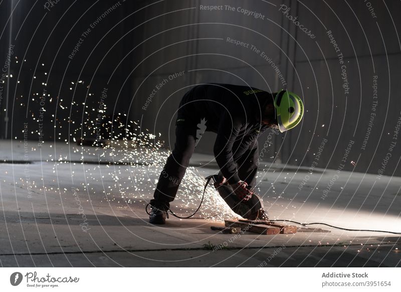 Anonymous male technician cutting metal with chop saw on street at night man welder iron spark metalwork manufacture tool industrial worker labor job helmet
