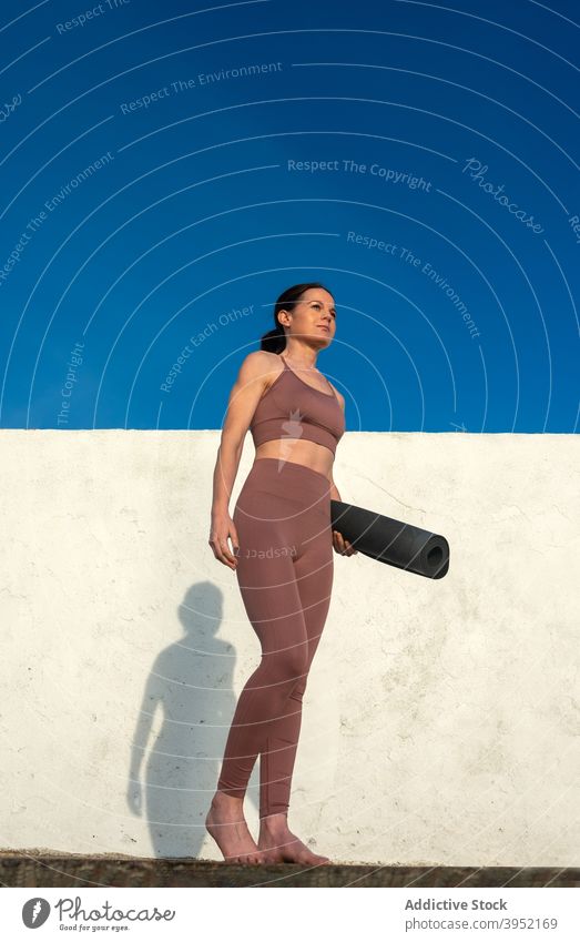 Tranquil woman with yoga mat standing near wall slim tranquil activewear ready prepare sportswear harmony female concrete sky blue sunny healthy slender serene