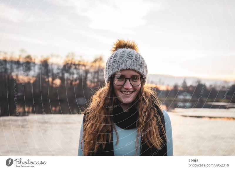 Smiling young woman with glasses and wool cap by a lake Woman youthful Winter portrait Lake Woolen hat Scarf long hair Curly Red pretty Autumn Nature Happy Cute