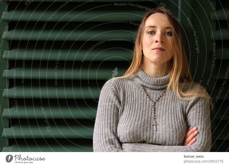 A woman with a narrow face wears a rosary around her neck and stands leaning against green blinds Woman Sweater Rosary Green green background Head portrait