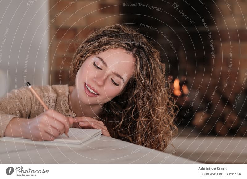 Smiling woman taking notes in notebook at home write diary fireplace cozy take note smile domestic female house content rest cottage comfort joy sit relax happy