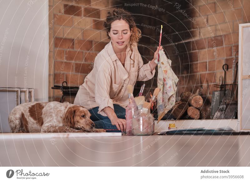 Cheerful painter and dog sitting on floor woman artist clean paintbrush setter together smile female owner messy house paper supply tool creative hobby painting