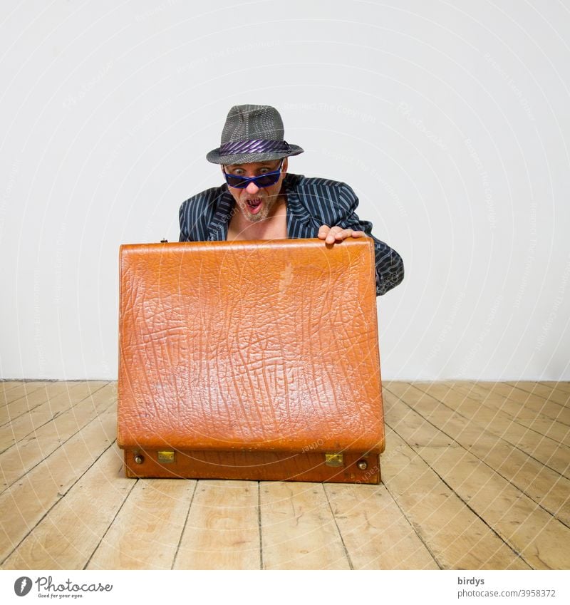 Man in suit with hat and sunglasses looks very surprised into an opened old suitcase. Surprise Suitcase astonishment Horror Looking Contents Wooden floor Marvel