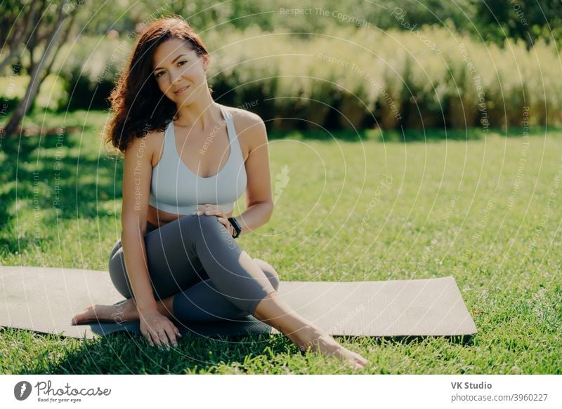 Photo of brunette sportswoman has rest after fitness training sits on karemat with bare feet in activewear leads healthy lifestyle poses on fresh green grass enjoys summy day and good weather