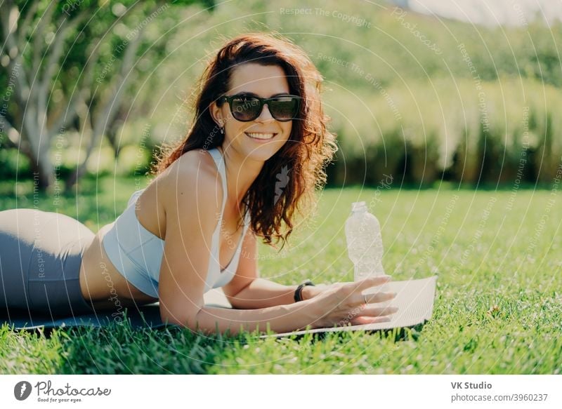 Smiling positive brunette woman lying at karemat on fitness mat wears sunglasses cropped top and leggings holds bottle of water enjoys sunny day breathes fresh air has rest after gymnastic exercises