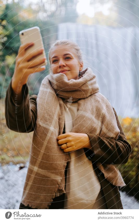 Smiling woman taking selfie in forest against waterfall smartphone autumn outerwear self portrait cheerful take photo female season amazing memory mobile moment