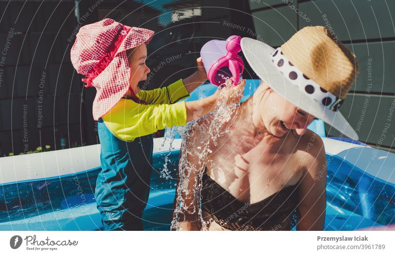 Baby pouring water on mother in baby swimming pool. summer child girl fun inflatable candid lifestyle authentic sun protection hat family motherhood happy warm