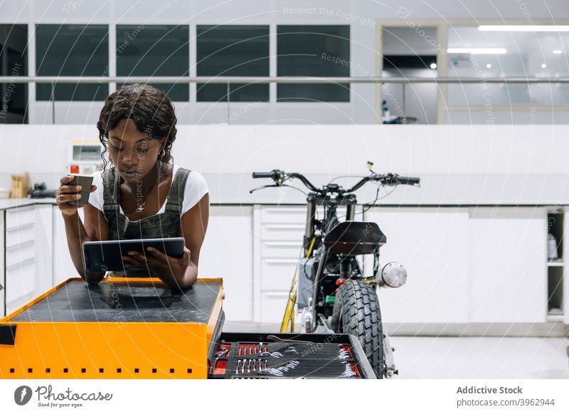 Black mechanic browsing tablet in motorbike workshop woman technician drink coffee female ethnic black african american uniform using job tool cabinet modern