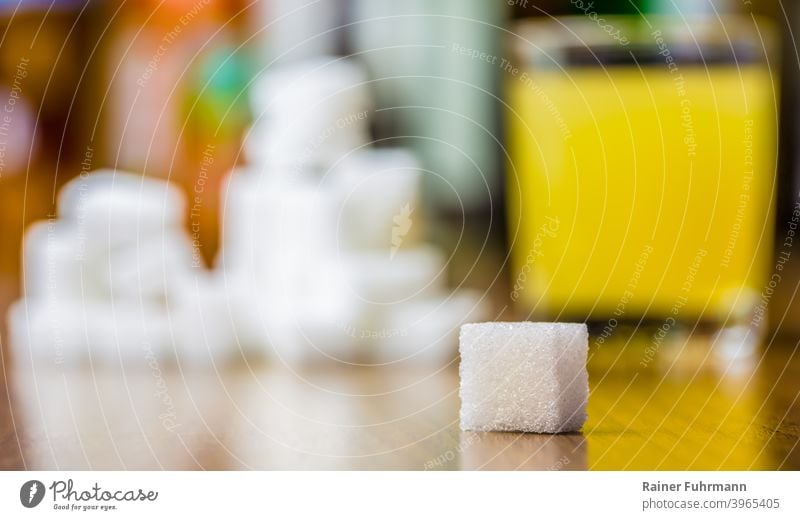 A piece of sugar cube is lying on a table. A glass with juice and many pieces of sugar are in the blurred background. Sugar Lump sugar Carbohydrates cute Food