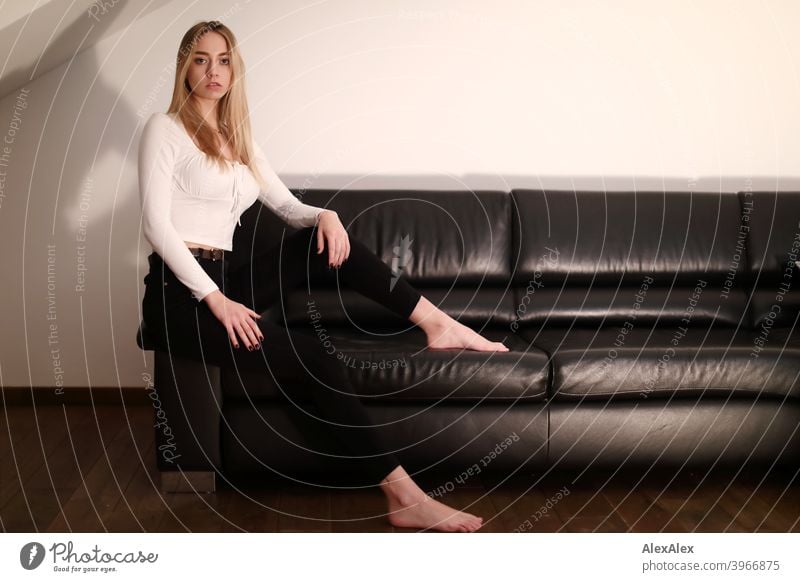 Portrait of a young blonde woman sitting barefoot on a black leather couch and watching Woman Girl Slim Blonde Long-haired Dark portrait Night pretty Graceful