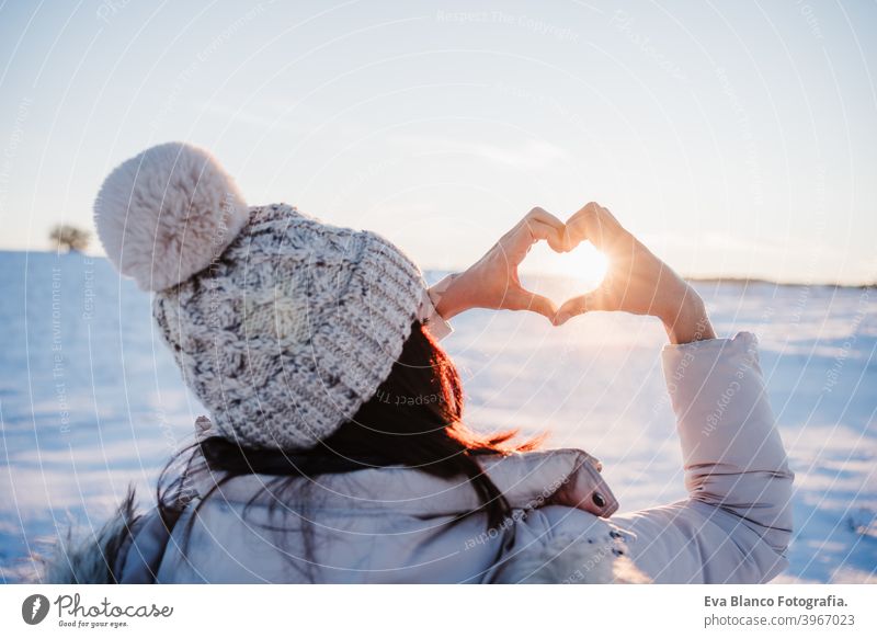 happy young woman hiking in snowy mountain at sunset doing a heart shape with hands. winter season. nature and love back view cold caucasian wanderlust travel