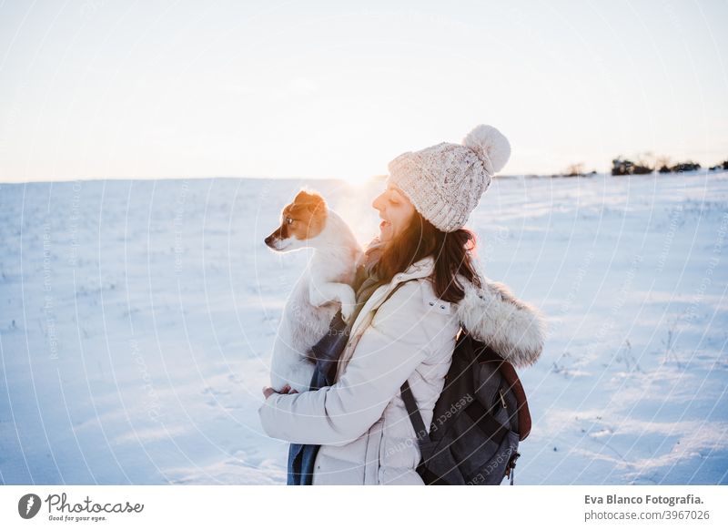 happy woman in snowy mountain wearing modern coat at sunset. Holding cute jack russell dog in arms. winter season. nature at sunset travel owner pet love