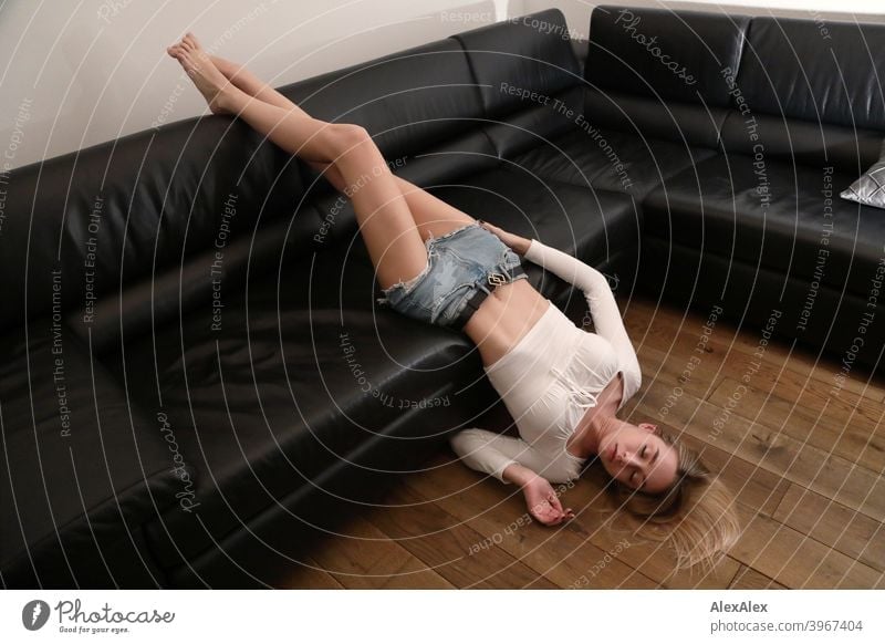 Portrait of a young blonde woman in hot pants lying barefoot and upside down on a black leather couch Woman Girl Slim Blonde Long-haired Dark portrait pretty