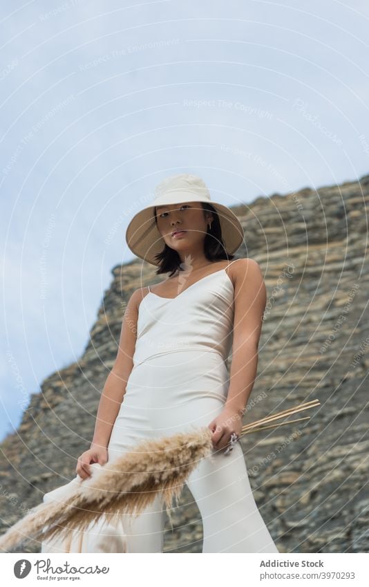 Stylish ethnic woman in hat standing near rock style fashion outfit white garment cloth apparel young asian female bouquet nature trendy elegant lady slim