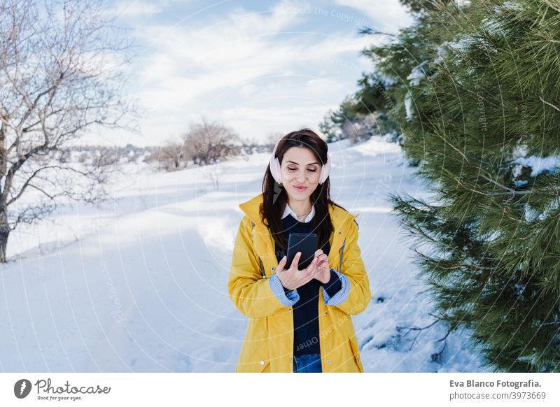 young beautiful woman listening to music on mobile phone and headset in the snow. wearing yellow coat, winter lifestyle mountain technology daytime caucasian