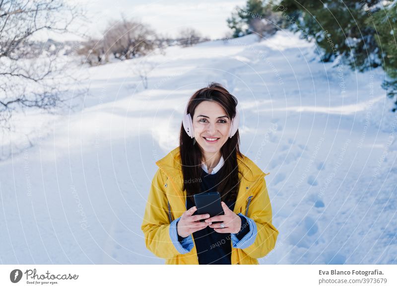young beautiful woman listening to music on mobile phone and headset in the snow. wearing yellow coat, winter lifestyle mountain technology daytime caucasian