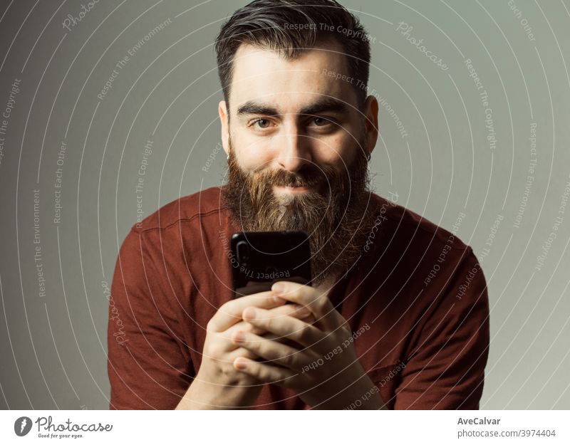 A young hipster style man with a beard and modern haircut smiling while looking at camera with his phone screen with copy space laughing colourful living
