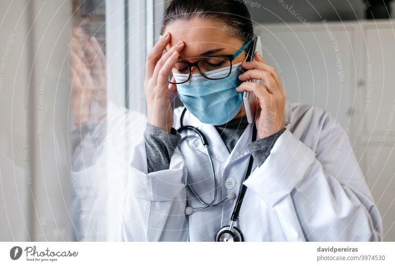 Worried female doctor talking on the phone mobile sad bad news pandemic fatigue worried stress desperate protective mask closed eyes hand on head coronavirus