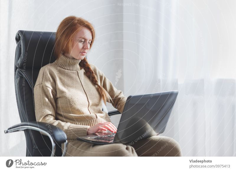 Young caucasian woman sitting in leather armchair using laptop working from home, freelance, social distancing and remote work concept. typing study redhead