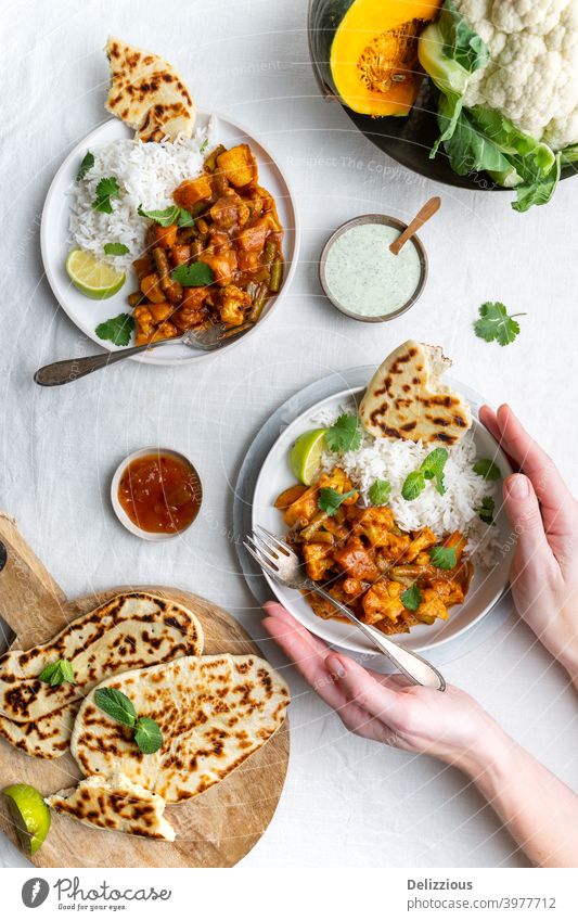 Top down view of a vegetarian, vegan Indian Balti curry with cauliflower and pumpkin served with raita, naan bread and mango chutney and rice, female hands holding one plate, white background