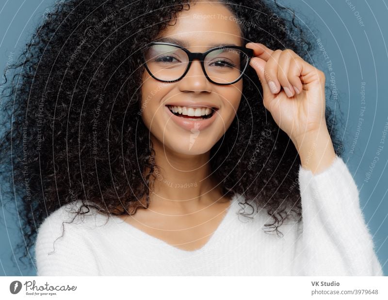 Cropped shot of good looking woman wears optical glasses, smiles broadly, shows white perfect teeth, healthy dark skin, dressed in casual jumper, isolated over blue background. Feminity concept