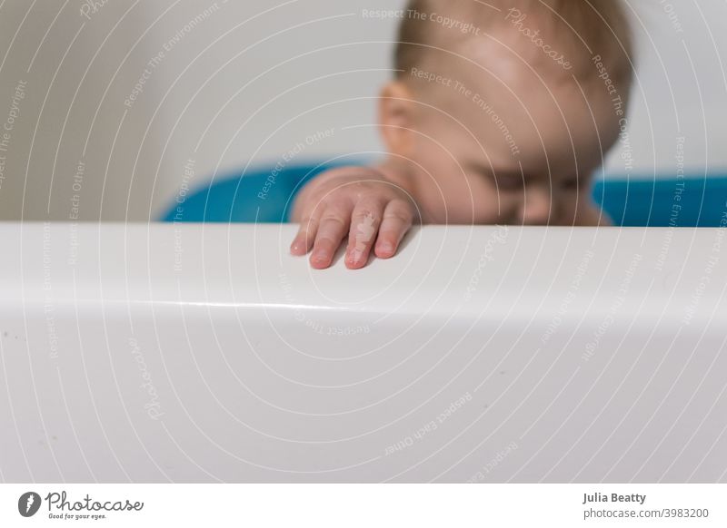 Close up of baby's hands and fingers grasping edge of bathtub; in background baby is sitting upright in blue infant bath tub bath time clean reach seated child