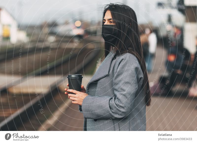 Happy girl in a mask stands waiting for the train at the station with coffee in her hands. transport young female beautiful railway passenger metro woman