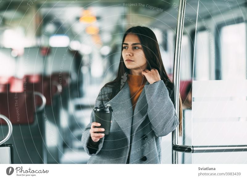 A happy brunette with coffee in her hands stands on the train, looks into the distance and smiles. transport young female beautiful railway passenger metro