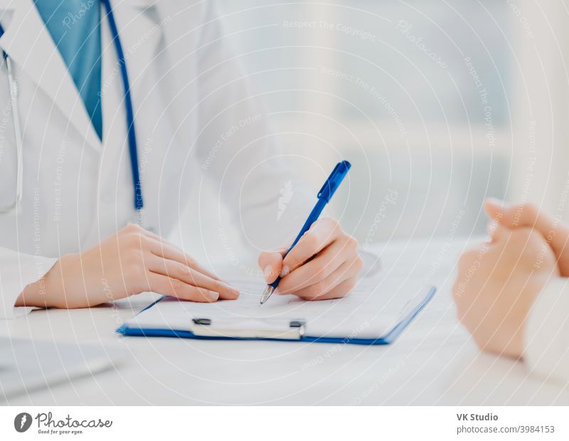 Unknown female doctor writes down prescription on papers in clipboard, consults patient about curing disease, pose at desktop against blurred white background fills up medical form. Symptoms diagnosis