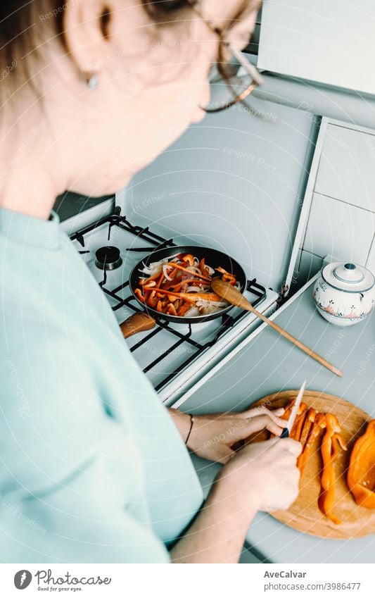 A woman cutting some pepper over a wooden round plate with a knife while cooking with the pan in the kitchen horizontal workplace desktop person indoors