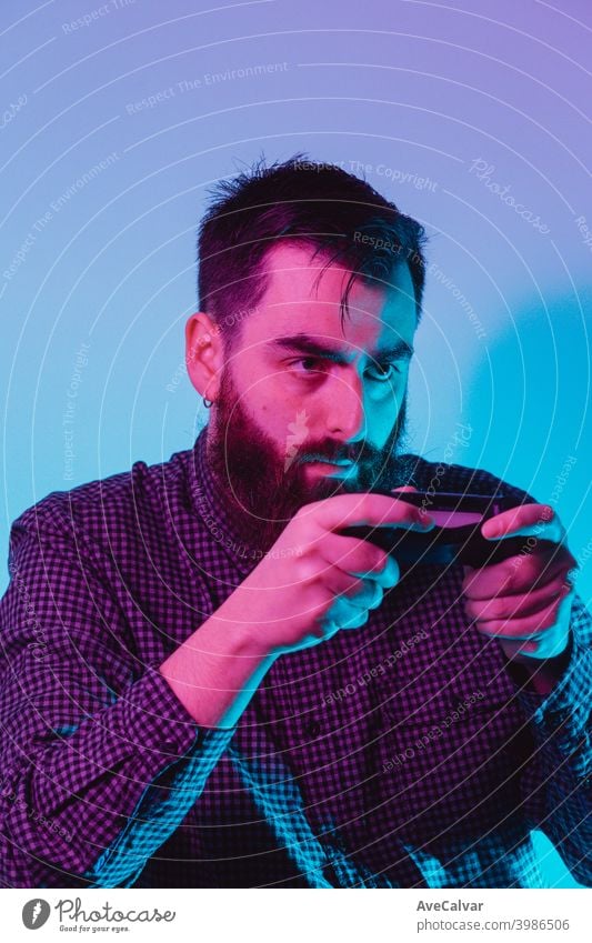 A young hipster man holding a console pad while playing with a serious face with pink and blue lights 20s copy-space exciting videogame technology expressive